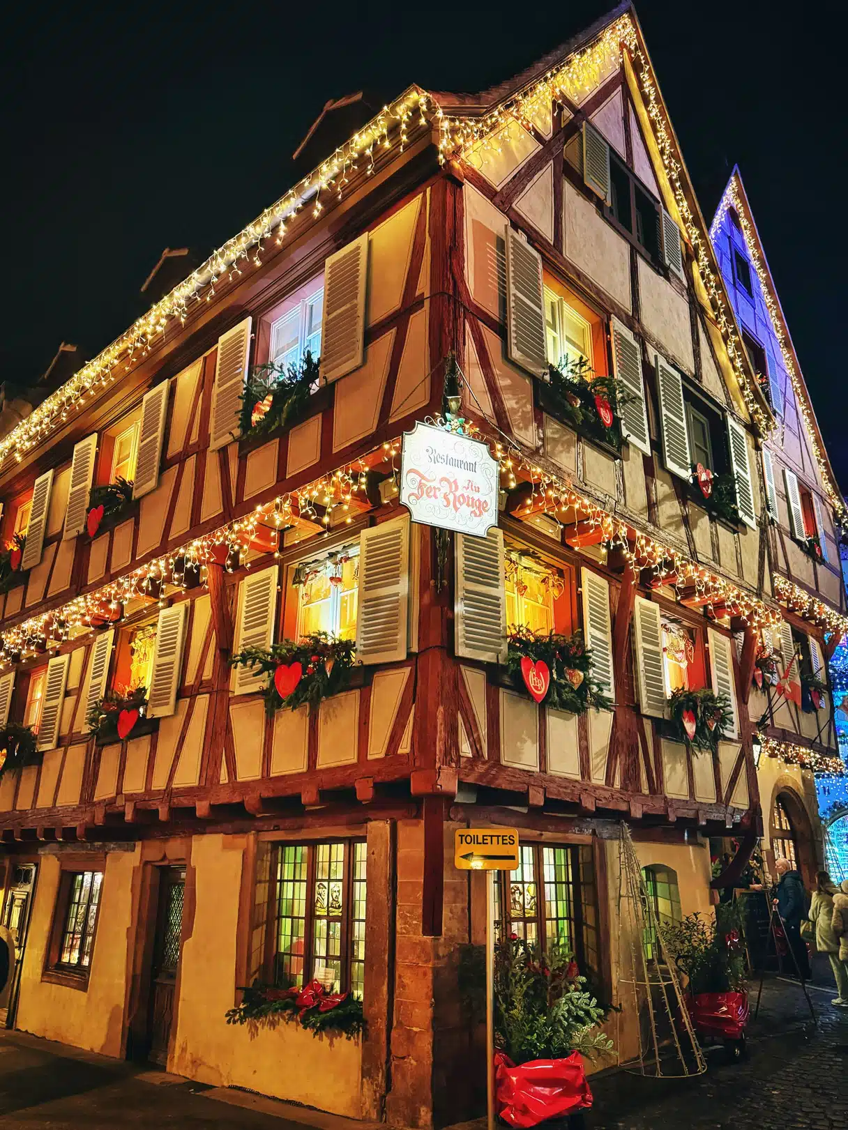 Colmar’da yarı ahşap Au Fer Rouge restoranı gece; cephesi Noel ışıklarıyla çevrelenmiş, köknar çelenkleri ve kırmızı kalplerle süslenmiş