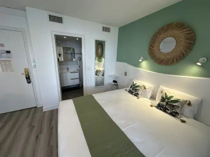 Yeşil vurgu duvarı, hasır çerçeveli aynası ve özel banyosu olan modern bir çift kişilik otel odası