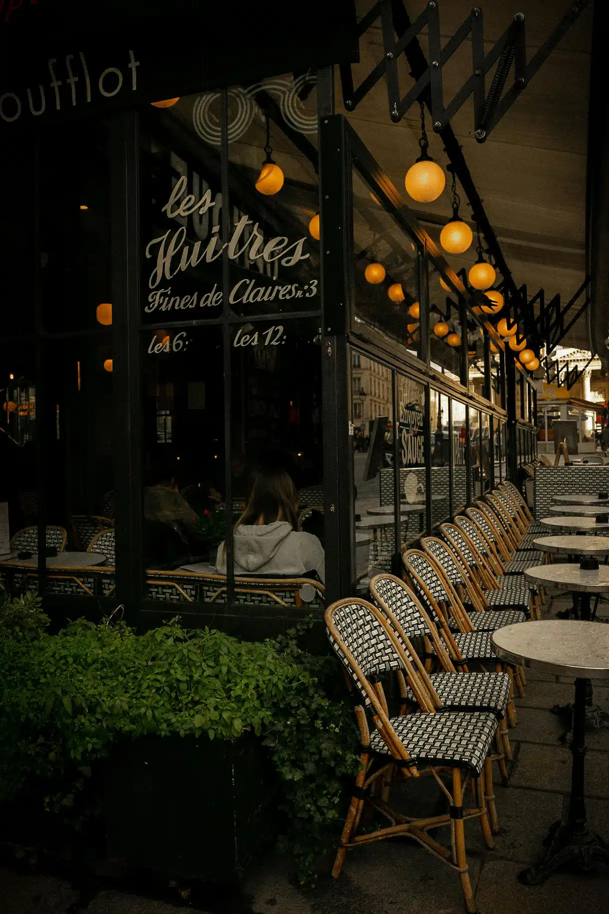 Empty rattan chairs on a Paris café terrace lit by glowing globe lights in the evening, with “Les Huîtres” painted on the window