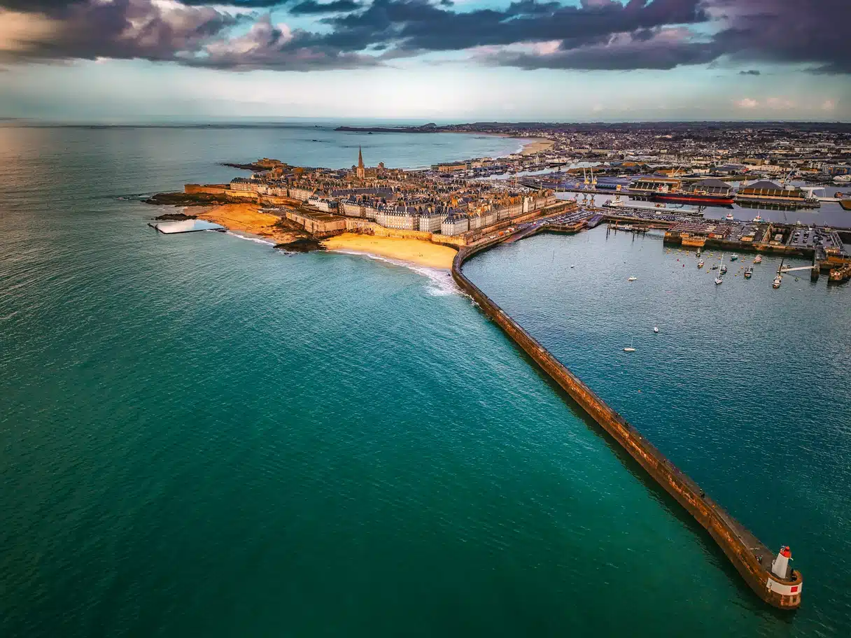 Aerial view of the walled city of Saint-Malo, its beach and the long harbour breakwater, the ramparts lit by warm evening light under a bank of dark cloud